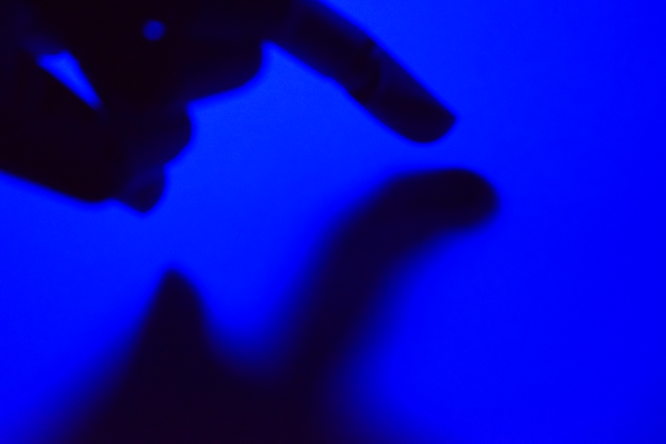 A close-up abstract image featuring a silhouette of a hand against a vibrant blue background. The blurred edges and interplay of light and shadow create a sense of mystery and depth, inviting interpretation and introspection. Captured on a Nikon D5300 digital camera. A work by John Paglia.