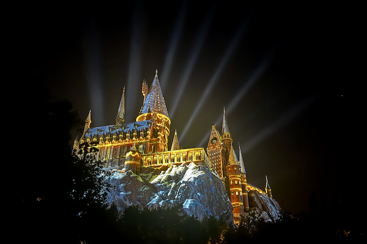 A stunning nighttime image of Hogwarts Castle from the Wizarding World of Harry Potter, located in Hogsmeade at Islands of Adventure in Universal Orlando Resort. The castle is illuminated with golden lights and snow-like effects on the rooftops, with dramatic beams of light radiating into the night sky. The intricate architecture, including spires and detailed stonework, captures the magical essence of the Harry Potter universe. Shot on iPhone. Shot on iPhone. A work by John Paglia.
