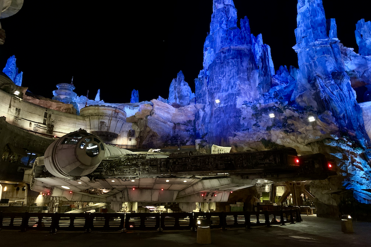 A nighttime image of the Millennium Falcon, the iconic spaceship from Star Wars, stationed at Galaxy's Edge in Disney's Hollywood Studios, Walt Disney World. The detailed replica is illuminated under soft white and red lighting, set against the backdrop of towering, otherworldly rock formations bathed in blue light, evoking the planetary setting of Batuu. The scene immerses viewers in the universe of Star Wars with its authentic design and dramatic lighting. Shot on iPhone. A work by John Paglia.