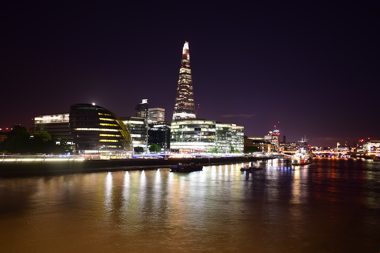 A stunning nighttime view of London's skyline featuring The Shard and surrounding modern architecture along the Thames River. The buildings are illuminated, reflecting vibrant lights on the calm water, while the HMS Belfast is visible docked nearby. Captured on a Nikon D5300 digital camera. A work by John Paglia.