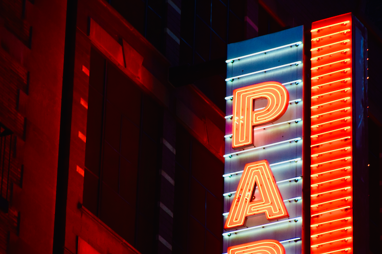 A close-up nighttime shot of a neon sign spelling “PARK,” glowing brightly in red and blue hues. The vibrant lights contrast against the deep shadows of the building façade, which is bathed in moody red lighting. The image captures the retro aesthetic and urban atmosphere of a city at night. Captured on a Nikon D5300 digital camera. A work by John Paglia.