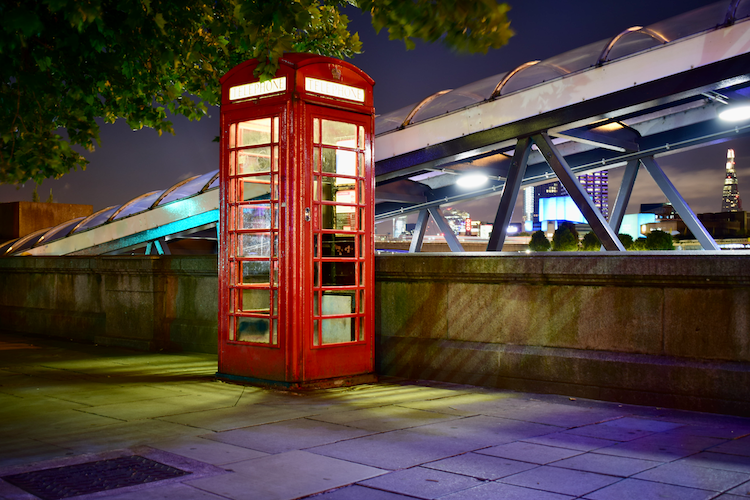 A classic red telephone booth illuminated against the night sky at Embankment, London. The vibrant structure stands near modern glass and steel architectural elements, with The Shard visible in the distant background. Soft, colorful lighting reflects off the pavement, adding a contemporary touch to the historic scene. Captured on a Nikon D5300 digital camera. A work by John Paglia.