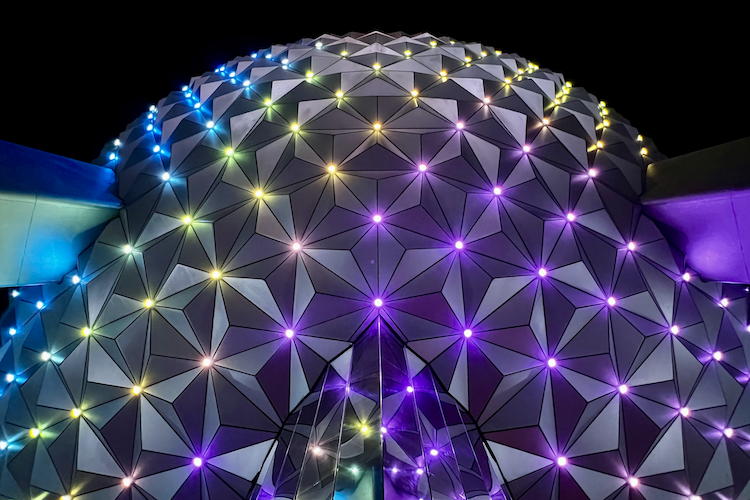 A close-up view of Spaceship Earth at Epcot in Walt Disney World illuminated at night, showcasing its iconic geodesic dome structure adorned with multicolored LED lights. The geometric panels form a mesmerizing pattern, with vibrant hues of purple, yellow, and blue creating a captivating display against the dark sky. Shot on iPhone. A work by John Paglia.