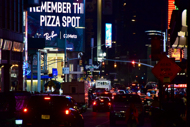 A bustling nighttime view of Times Square, New York City, with glowing neon lights and illuminated advertisements, including a prominent “Remember This Pizza Spot” billboard. The street is crowded with cars, taxis, and pedestrians, capturing the vibrant energy of the iconic location. Captured on a Nikon D5300 digital camera. A work by John Paglia.