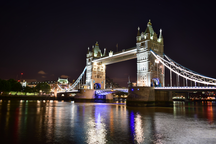 A stunning nighttime photograph of Tower Bridge in London, illuminated with vibrant white and blue lights. The iconic suspension bridge, with its twin gothic-style towers, reflects beautifully on the calm waters of the River Thames below. The scene captures the architectural grandeur of the bridge, surrounded by the soft glow of the city's lights, creating a serene and captivating ambiance. Captured on a Nikon D5300 digital camera. A work by John Paglia.