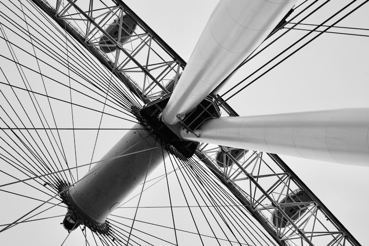 A close-up black-and-white photograph of the London Eye, capturing the intricate network of steel cables and the central hub of the giant observation wheel. The perspective emphasizes the architectural precision and engineering brilliance of this iconic structure. The repeating geometric patterns create a striking and dynamic composition against the light background. Captured on a Nikon D5300 digital camera. A work by John Paglia.