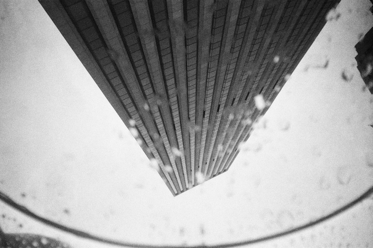 A black-and-white photograph of 767 Fifth Avenue in New York City, reflected through a skylight at the Apple Store below the square. The image captures the building's towering structure, distorted slightly by the curved reflection, with soft light and subtle water droplets adding texture and depth to the composition. Captured on an Argus C3 camera with Ilford HP5 Plus film. A work by John Paglia.