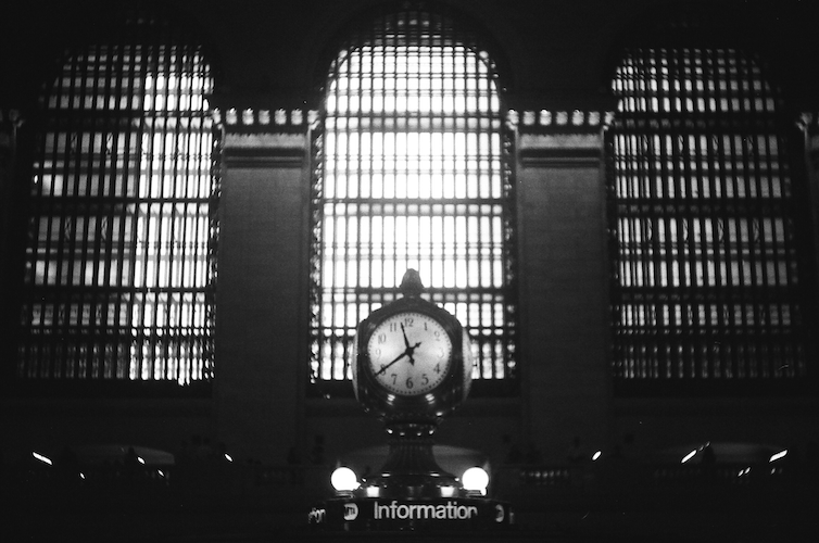 A black-and-white photograph of the iconic clock inside Grand Central Terminal, New York City. The image highlights the ornate timepiece above the information booth, framed by the station's towering arched windows with intricate gridded patterns. The contrast of light streaming through the windows emphasizes the grandeur of this historic transportation hub. This timeless landmark symbolizes New York's architectural and cultural heritage. Captured on an Argus C3 with Ilford HP5 Plus film. - A Work by John Paglia