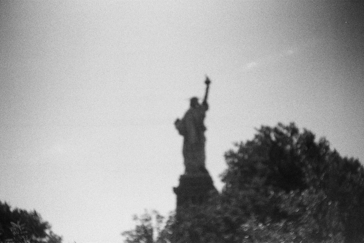 A black-and-white photograph of the Statue of Liberty, partially obscured by trees in the foreground. The iconic figure is slightly out of focus, creating a dreamlike quality that emphasizes its silhouette against the expansive sky. The image captures the timeless essence of this global symbol of freedom and hope. Captured on an Argus C3 camera with Ilford HP5 Plus film. A work by John Paglia.