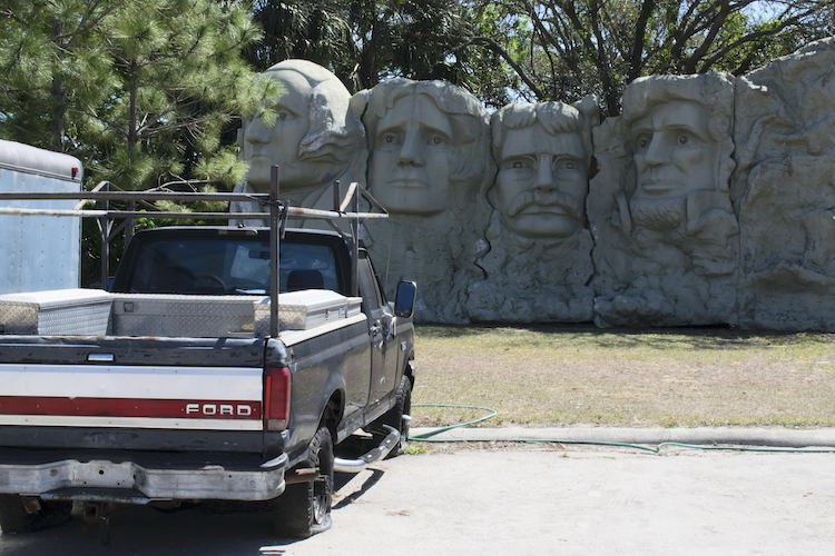 A black Ford pickup truck is parked in front of a sculptural wall featuring four large stone faces resembling Mount Rushmore but with exaggerated features. The scene is set outdoors with trees and a trailer visible in the background. Captured on a Nikon D5300 digital camera. A work by John Paglia.
