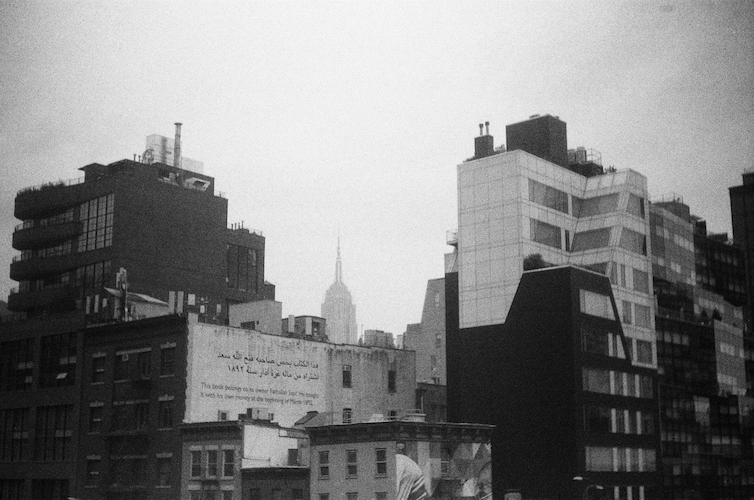 A black-and-white cityscape capturing modern and vintage architecture in New York City. The Empire State Building rises subtly in the hazy background, creating a moody, timeless atmosphere. Ideal for themes of urban exploration, cinematic photography, and architectural contrast. Captured on an Argus C3 camera with Ilford HP5 Plus film. A work by John Paglia.