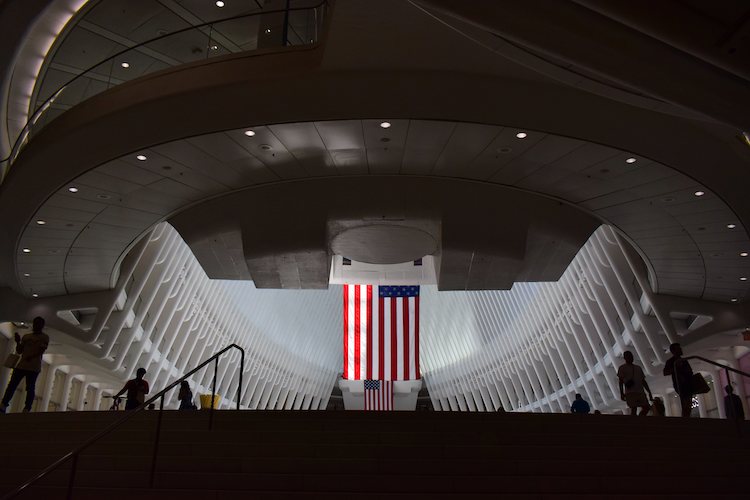 A striking view of the Oculus in New York City, capturing the dramatic architecture of the transportation hub. The image features the soaring white rib-like structures framing a large American flag in the center. Silhouettes of people walking up the stairs add a sense of scale to the grand, futuristic design. The soft lighting highlights the modern elegance of the space. Captured on a Nikon D5300 digital camera. A work by John Paglia.