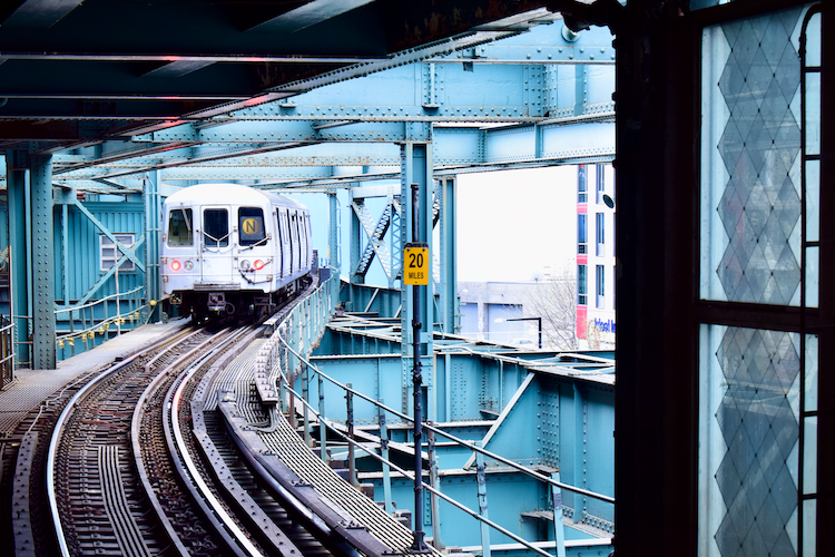 Queensboro Plaza Station in NYC, with an N train pulling away under teal steel beams, capturing urban transit at dusk.