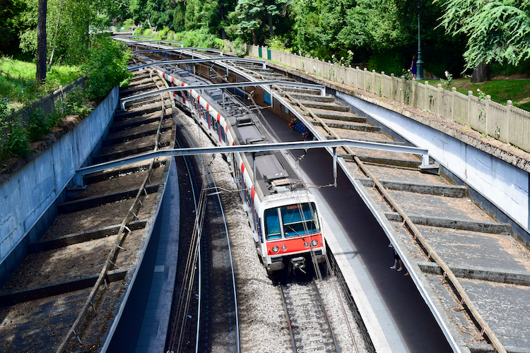 A vibrant scene at Cité Universitaire station in Paris, showcasing a red and white RER B train arriving at the platform. The station's tracks are flanked by metallic structures and bordered by lush greenery, creating a blend of urban design and natural beauty. The overhead wires and concrete details emphasize the functionality of this transit hub. Captured on a Nikon D5300 digital camera. A work by John Paglia.