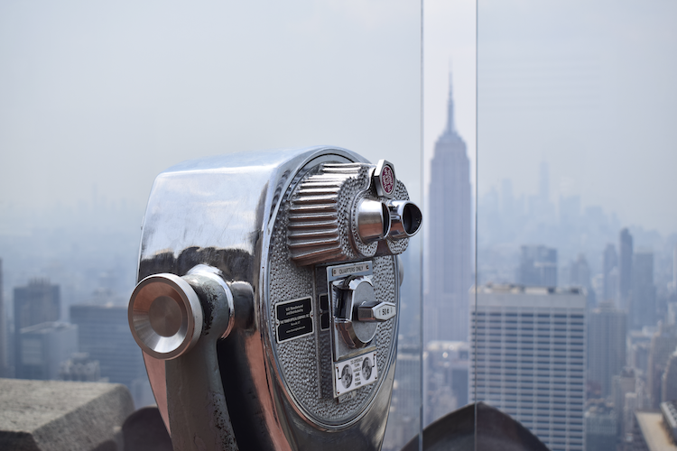 A coin-operated binocular viewer at Top of the Rock in New York City, with the Empire State Building visible in the hazy skyline. The chrome details of the viewer contrast with the soft, muted tones of the urban landscape, creating a classic perspective of the city's iconic architecture. Captured on a Nikon D5300 digital camera. A work by John Paglia.