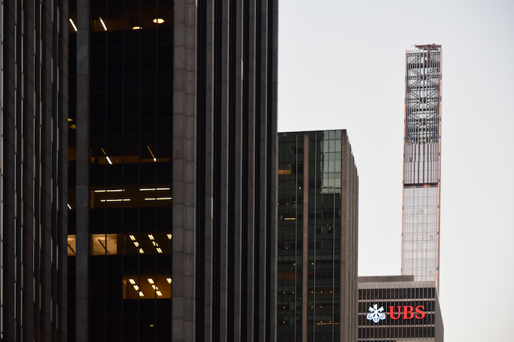 A view of Sixth Avenue in New York City, featuring a mix of modern skyscrapers and office buildings. The UBS logo is prominently displayed on one of the structures, while the distinctive slender design of 111 West 57th Street, also known as the Steinway Tower, stands tall in the background. The contrast between illuminated office windows and the fading daylight creates a dynamic urban atmosphere. Captured on a Nikon D5300 digital camera. A work by John Paglia.