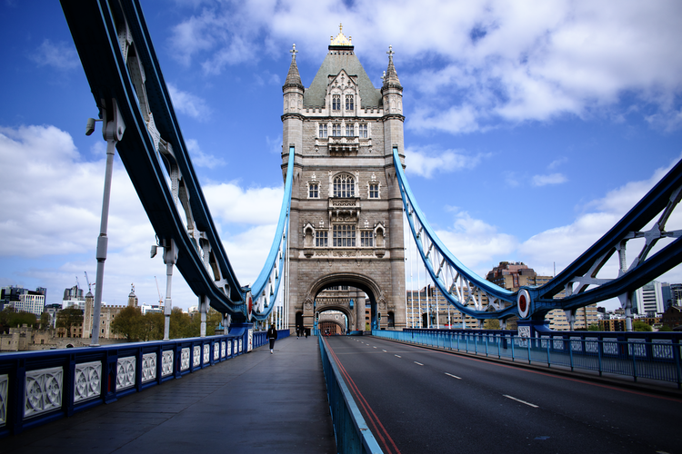 A photograph of Tower Bridge in London taken during the COVID-19 lockdown in 2020. The iconic structure stands against a bright blue sky with minimal traffic, creating an eerily quiet scene. The bridge's detailed architecture and vibrant blue suspension cables are prominently featured, contrasting with the empty road and pedestrian walkway. The surrounding cityscape adds context to this historic moment. Captured on a Nikon D5300 digital camera. A work by John Paglia.