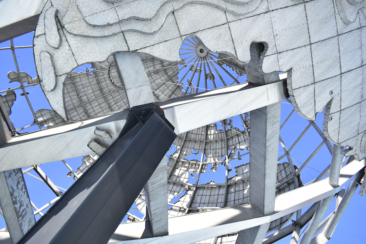 A close-up view of the Unisphere in Flushing Meadows-Corona Park, New York City, showcasing its intricate stainless steel design. The structure's grid-like representation of continents and supporting beams is highlighted against a bright blue sky, emphasizing its iconic presence. The geometric composition captures the grandeur and detail of this celebrated landmark. Captured on a Nikon D5300 digital camera. A work by John Paglia.