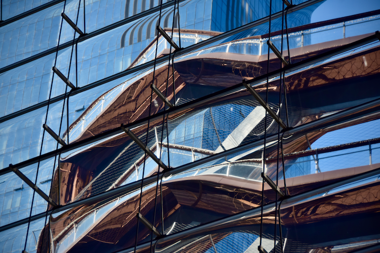 A view of The Vessel at Hudson Yards, designed by Thomas Heatherwick, seen through a window at The Shops at 30 Hudson Yards. The copper-colored geometric structure stands prominently, framed by the window's metal supports. The intricate design of The Vessel contrasts with the surrounding blue sky and modern architecture, creating a striking and layered perspective. Captured on a Nikon D5300 digital camera. A work by John Paglia.
