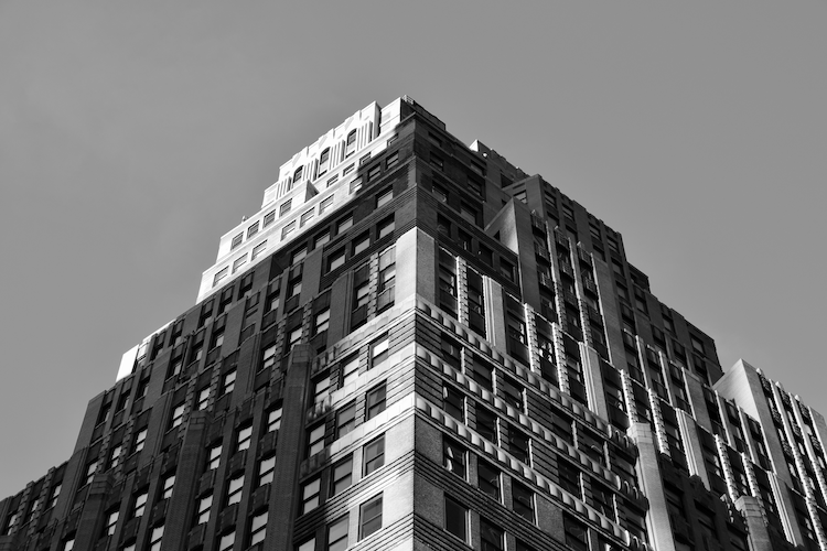 A black-and-white photograph of the building at 1441 Broadway in New York City, showcasing its iconic Art Deco architecture. The image highlights the intricate geometric patterns, brickwork, and tiered facade of the skyscraper, with sunlight and shadow creating a dynamic play across its surface. The upward perspective emphasizes the building's towering presence against a minimal sky backdrop. Captured on a Nikon D5300 digital camera. A work by John Paglia.