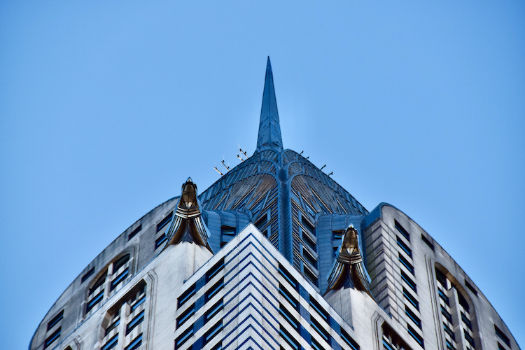 Close-up of the Chrysler Building's iconic Art Deco crown in New York City, highlighting its intricate geometric patterns, gleaming metallic details, and sharp spire against a vibrant blue sky. This architectural masterpiece exemplifies 1930s design and modern urban elegance. Captured on a Nikon D5300 digital camera. A work by John Paglia.