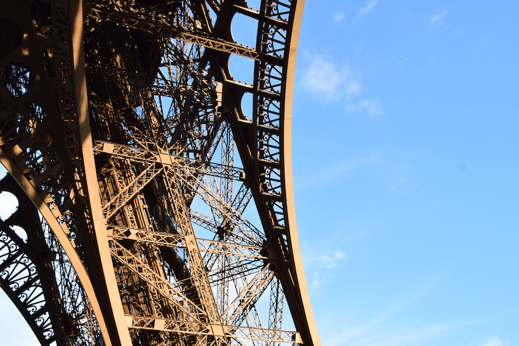 A detailed view of the intricate iron lattice structure of the Eiffel Tower in Paris, France. The warm sunlight highlights the elegant curves and patterns of the iconic architectural masterpiece against a bright blue sky, emphasizing its grandeur and craftsmanship. Captured on a Nikon D5300 digital camera. A work by John Paglia.