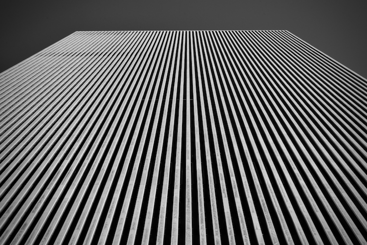 A black-and-white image showcasing the towering facade of the former Exxon Building in New York City. The perspective looks straight up, emphasizing the building's uniform vertical lines and geometric precision. The repetitive pattern of the sleek aluminum facade creates a striking sense of height and modernist design, set against a stark sky for dramatic contrast. Captured on a Nikon D5300 digital camera. A work by John Paglia.