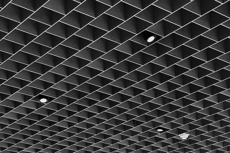 A black and white photograph of the geometric ceiling at the Museum of Modern Art (MoMA) in New York City. The ceiling features a grid of square recesses, creating a striking pattern with sharp contrasts and symmetry. Integrated lighting fixtures punctuate the design, adding a modern and minimalist aesthetic. Shot on iPhone. A Work by John Paglia.