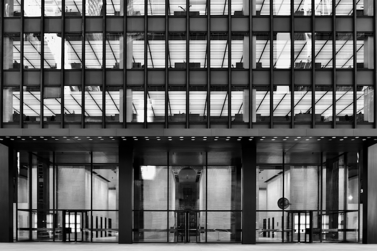 A black-and-white photograph of the iconic Seagram Building in New York City, showcasing its minimalist architectural design. The facade features a grid of rectangular windows framed by clean, dark lines, reflecting the International Style. The ground floor displays tall glass panels, offering a glimpse into the building's lobby with its understated elegance and symmetry. The photograph emphasizes the structure's geometric precision and modernist aesthetic. Captured on a Nikon D5300 digital camera. A work by John Paglia.