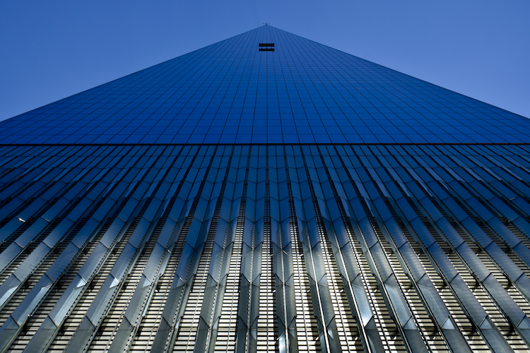 A striking upward view of One World Trade Center in New York City, emphasizing its geometric facade and sleek, modern design. The building's reflective glass panels and tapered structure create a visually dynamic effect as the lines converge toward the pinnacle. The sunlight highlights the intricate patterns of the lower metal fins, contrasting with the deep blue sky in the background, showcasing the architectural brilliance of this iconic skyscraper. Captured on a Nikon D5300 digital camera. A work by John Paglia.