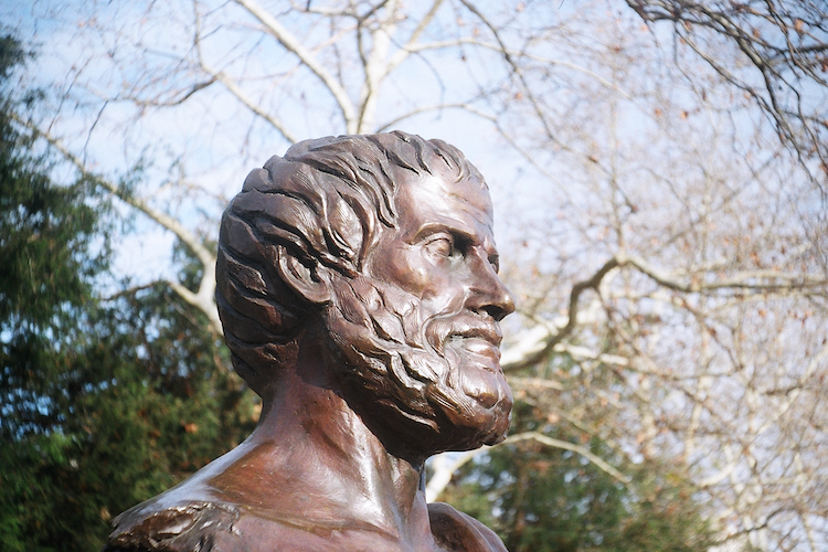 A bronze statue of Aristotle located in Athens Square Park in Astoria, Queens. The statue captures the philosopher's profile, with his detailed beard and contemplative expression highlighted against a backdrop of bare tree branches and a clear sky. The natural surroundings complement the timeless and reflective aura of the sculpture, paying homage to the iconic Greek thinker. Captured on an Argus C3 camera with Kodak Ektar film - A work by John Paglia