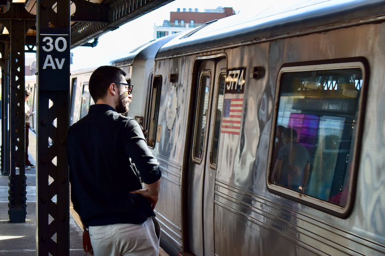 A man stands on the 30th Avenue subway platform in Astoria, Queens, watching as a silver train pulls into the station. Dressed in a black shirt and light-colored pants, he leans slightly against a metal column with the station's name sign visible. The reflective surface of the train captures blurred glimpses of passengers and vibrant colors. The urban background, with hints of buildings and sunlight, adds depth to the dynamic scene of daily transit life in New York City. Captured on a Nikon D5300 digital camera. - A work by John Paglia