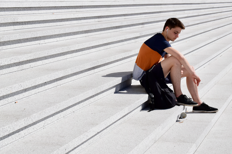 A young man sitting on the stairs of La Grande Arche de la Défense in Paris, wearing a color-blocked orange and navy t-shirt, black shorts, and black sneakers. He has white earbuds in and appears lost in thought, with a skateboard resting nearby on the steps. The minimalist architecture and the sharp lines of the stone steps frame the serene and contemplative moment. Captured on a Nikon D5300 digital camera. - A work by John Paglia