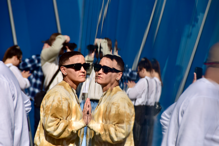 A striking photograph taken at The Edge at 30 Hudson Yards, featuring a person wearing a tie-dye yellow sweatshirt, black sunglasses, and gold jewelry. They are leaning against a reflective glass panel, creating a sharp mirror image that adds depth and symmetry to the composition. In the background, blurred figures of visitors engage with the space, contrasting against the deep blue architectural elements. The reflection emphasizes the sleek and modern design of the iconic observation deck. Captured on a Nikon D5300 digital camera. - A work by John Paglia