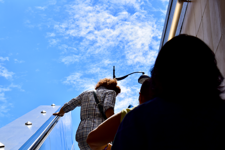 A vibrant scene at Luxembourg Station in Paris, capturing the upward view of commuters ascending an outdoor staircase. A person in the foreground wears a checkered outfit, their hand resting on the sleek metal railing, while other figures are silhouetted against the bright blue sky scattered with soft clouds. The composition highlights the architectural elements of the station and the dynamic transition from underground to open air. A streetlamp frames the scene, adding a Parisian touch to the vibrant moment. Captured on a Nikon D5300 digital camera. - A work by John Paglia