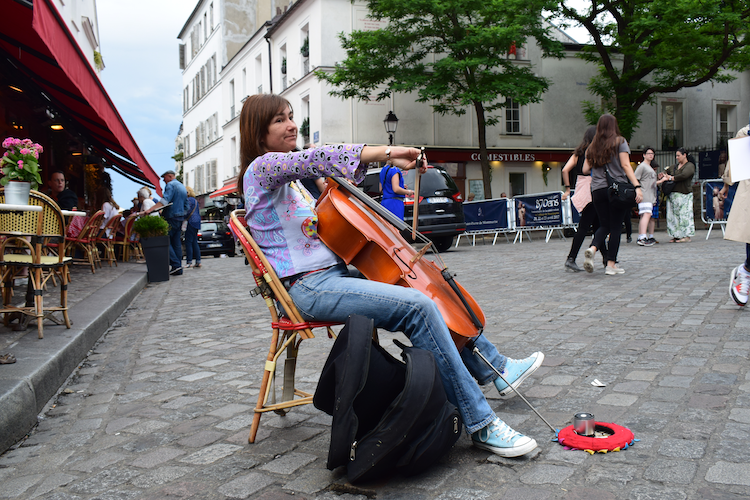 A street musician in Montmartre, Paris, plays the cello while seated on a red bistro chair on a cobblestone street. She wears a colorful patterned top, jeans, and light blue sneakers, with a cello case resting nearby. A small red mat with a cup for tips is placed on the ground in front of her. Behind her, a bustling scene unfolds with pedestrians, a tree-lined street, and charming Parisian cafés adorned with red awnings and outdoor seating, capturing the lively and artistic atmosphere of the area. Captured on a Nikon D5300 digital camera. - A work by John Paglia