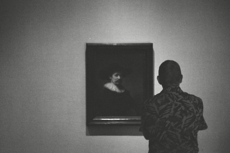 A black-and-white photograph of a visitor at The Met viewing Rembrandt van Rijn's portrait of Herman Doomer (ca. 1595–1650). The visitor, seen from behind, wears a patterned shirt and stands in quiet contemplation before the dark and expressive painting. The minimal gallery setting, with soft lighting focusing on the artwork, emphasizes the timelessness of Rembrandt's masterful depiction of texture and character. Captured on an Argus C3 camera with Ilford HP5 Plus film - A work by John Paglia