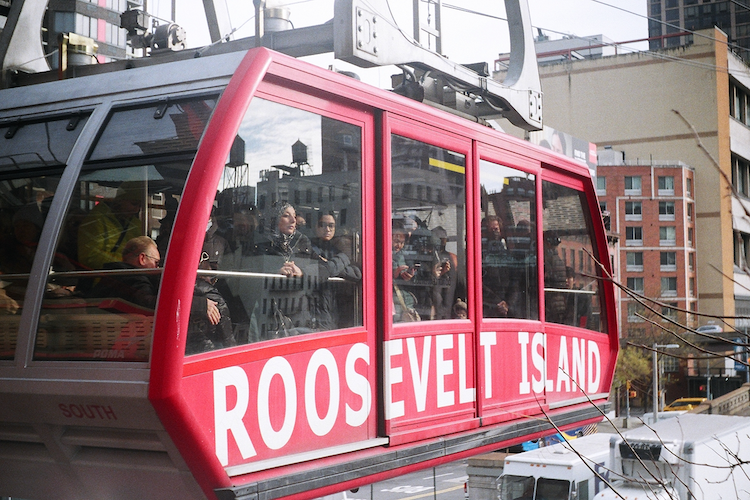A close-up photograph of the Roosevelt Island Tramway in New York City. The bright red tram car features large white text reading “ROOSEVELT ISLAND” and is filled with passengers enjoying the view. The background showcases urban buildings, including iconic water towers, under a partly cloudy sky. Captured on an Argus C3 with Kodak Ektar 100 film. - A Work by John Paglia