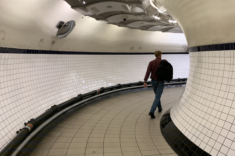 A curved tiled corridor at Shepherd's Bush Underground Station in London, showcasing the sleek white tiles with navy accents lining the walls and floor. A man wearing a flat cap and carrying a black backpack walks through the tunnel, emphasizing the dynamic movement and perspective of the space. The overhead lighting and exposed black cables along the wall add a modern, urban touch to the scene. Shot on iPhone. - A Work by John Paglia