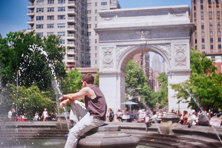 A lively summer scene in Washington Square Park, New York, where people gather near the grand arch.