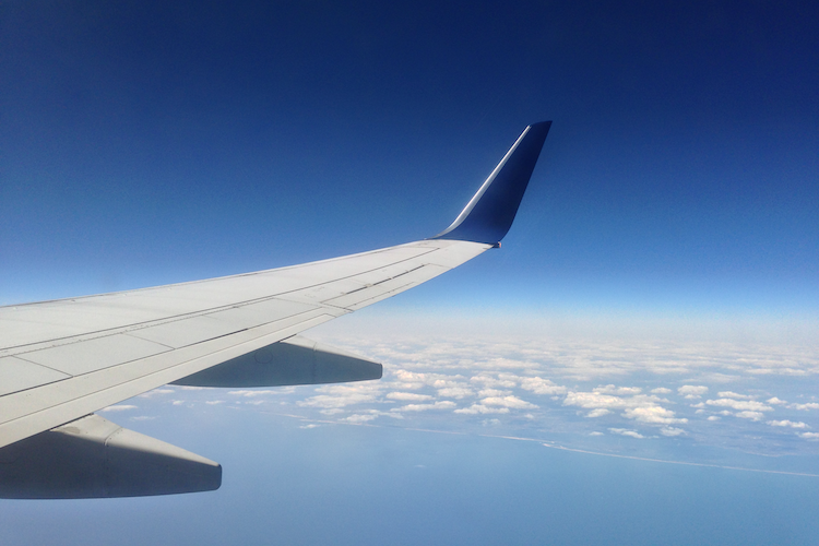 An aerial photograph capturing the view of an airplane wing extending into the clear blue sky. Beneath the wing, white clouds float over a serene stretch of water and a distant coastline, showcasing the beauty of flight and the vastness of the sky. Shot on iPhone. - A Work By John Paglia