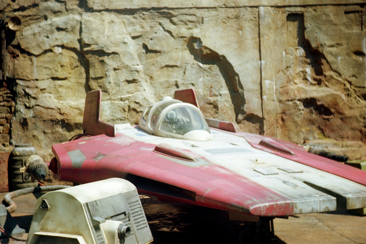 A detailed view of an A-wing starfighter from Star Wars: Galaxy's Edge in Disney's Hollywood Studios at Walt Disney World. The red and white A-wing is displayed against a weathered stone wall, evoking the rugged aesthetic of Batuu. Photographed with an Argus C3 camera on Kodak Ektar film, the image highlights the vibrant yet vintage tones of the starfighter's design, capturing the immersive details of the theme park's Star Wars-themed land. Captured on an Argus C3 camera with Kodak Ektar film. A Work by John Paglia.