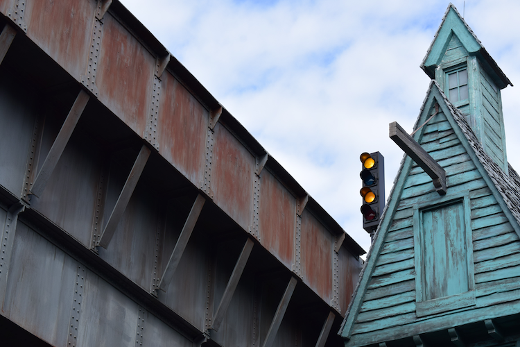 A detailed architectural view from Diagon Alley at Universal Studios Florida in Orlando, showcasing the contrast between the weathered turquoise facade of a whimsical, wizard-themed building and the rusty, industrial beams of a bridge structure. A traffic light with glowing yellow adds a touch of urban character to the magical atmosphere. The intricate textures and colors against the partly cloudy sky enhance the immersive design of the Wizarding World of Harry Potter. Captured on a Nikon D5300 digital camera. A Work by John Paglia.