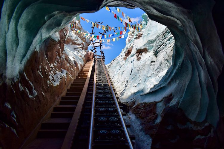 Expedition Everest roller coaster photography at Disney’s Animal Kingdom, showing the steep lift hill from inside a cavern with icy rock walls. The steel track and wooden staircase rise dramatically toward the sky, framed by snow-capped peaks and Tibetan prayer flags. Cinematic theme park photography capturing thrill, movement, and architectural detail in Orlando, Florida. Captured on a Nikon D5300 digital camera. A Work by John Paglia.