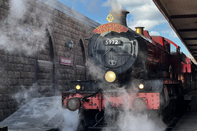 The Hogwarts Express arriving at Hogsmeade Station in Universal's Islands of Adventure, featuring its iconic red locomotive with “Hogwarts Express” written above the engine's headlamp. Steam rises dramatically around the train, adding an immersive and magical atmosphere. The background showcases the stone architecture of the Hogsmeade Station with its arched windows and the station sign. A vivid blue sky enhances the overall enchanting scene. Shot on iPhone. A Work by John Paglia.