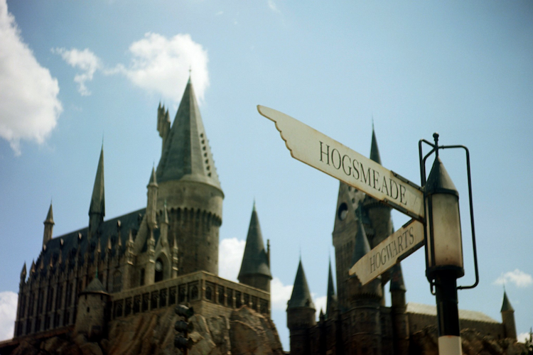 A magical view of Hogsmeade at Universal's Wizarding World of Harry Potter, Islands of Adventure. The iconic Hogwarts Castle towers in the background under a bright blue sky, with a vintage-style Hogsmeade directional sign in the foreground pointing toward Hogwarts. This photograph, taken on an Argus C3 camera with Kodak Ektar film, captures the enchanting atmosphere and nostalgic film tones, immersing viewers in the wizarding world. Captured on an Argus C3 camera with Kodak Ektar film. A Work by John Paglia.