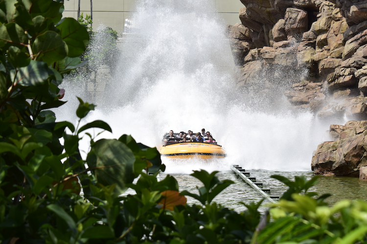 A thrilling moment captured on the Jurassic Park River Adventure ride at Universal's Islands of Adventure. The yellow raft plunges down the final drop, creating an enormous splash that drenches riders and spectators alike. Framed by lush green foliage in the foreground, the image highlights the ride's immersive dinosaur-themed setting, surrounded by rugged rock formations and cascading water. The exhilarated expressions of the riders emphasize the ride's high-energy excitement. Captured on a Nikon D5300 digital camera. A Work by John Paglia.