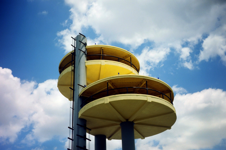 A close-up of the towering structures from the Men in Black: Alien Attack ride at Universal Studios Florida. The futuristic yellow platforms are supported by sleek columns and set against a bright blue sky with scattered white clouds. These elements mirror the ride's retro sci-fi aesthetic, reminiscent of mid-century design. The photograph captures the ride's iconic architecture, evoking the theme of alien adventures. Taken on an Argus C3 camera with Kodak Ektar film, the image boasts a nostalgic filmic charm. Captured on an Argus C3 camera with Kodak Ektar film. A Work by John Paglia.