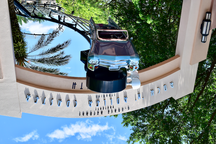 The iconic entrance sign for the Rock ‘n' Roller Coaster at Disney's Hollywood Studios in Walt Disney World, featuring a life-sized model of a classic convertible car suspended upside down above the archway. The surrounding vibrant green trees and a clear blue sky enhance the colorful details of the sign. The stylized musical roller coaster track loops into the background, symbolizing the thrilling ride's theme. Captured on a Nikon D5300 digital camera. A Work by John Paglia.