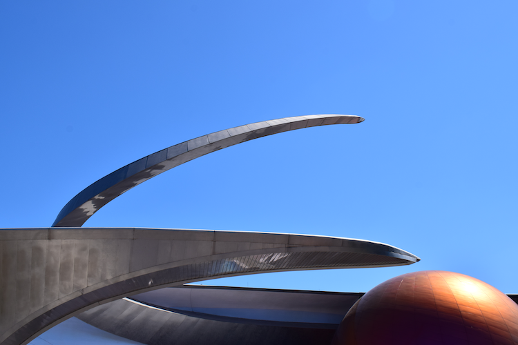 A striking architectural detail of the entrance to Mission: SPACE at Epcot, Walt Disney World. The photograph captures sleek, metallic curves juxtaposed with a vibrant orange planetary sphere against a clear, cloudless blue sky. The futuristic design of the structure emphasizes the theme of space exploration, inviting visitors to embark on an interstellar journey. Captured on a Nikon D5300 digital camera. A Work by John Paglia.